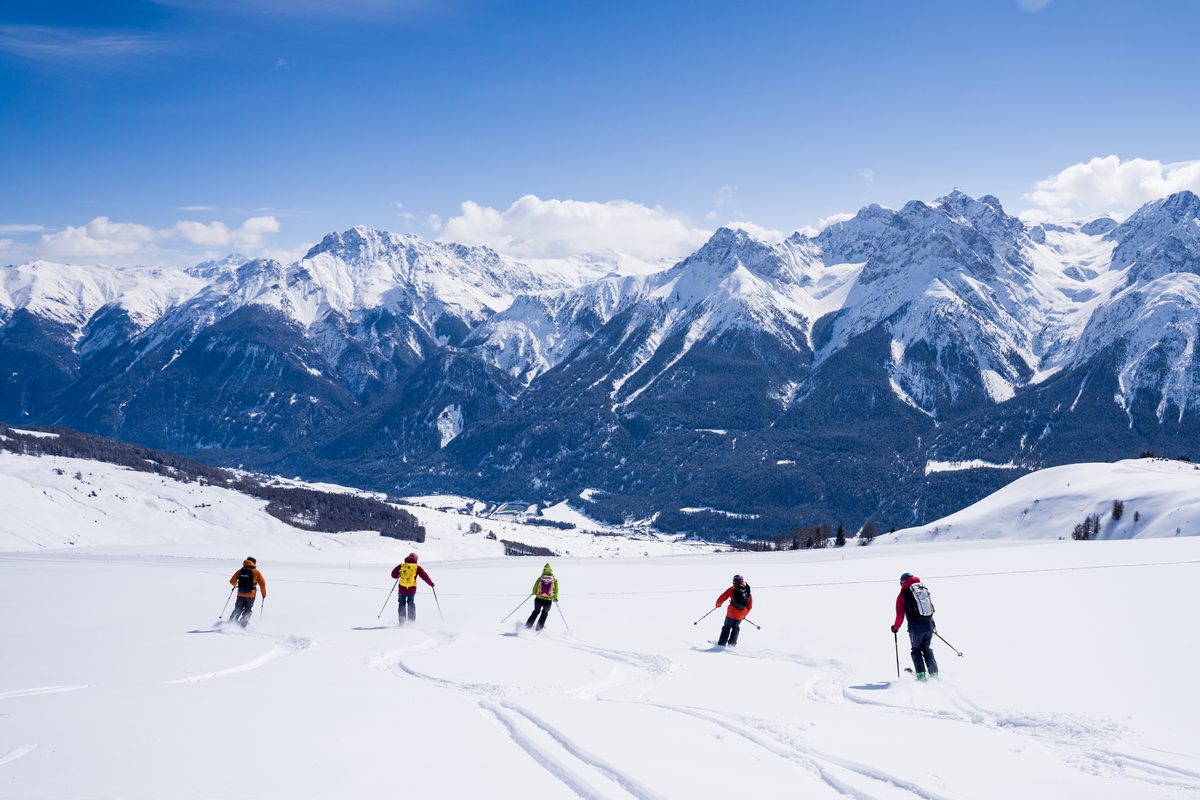 Skifahrende fahren einen breiten, verschneiten Hang hinunter, mit einer Reihe verschneiter Gipfel unter blauem Himmel.