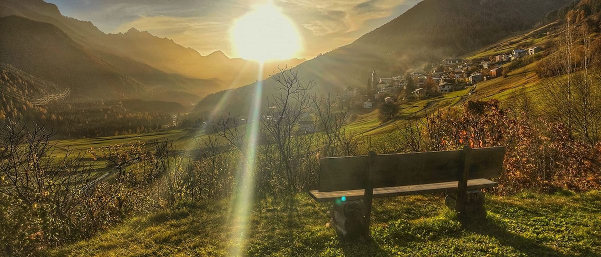 Eine leere Holzbank an einem Berghang, die Sonne tief über dem Tal und die Berge dahinter.
