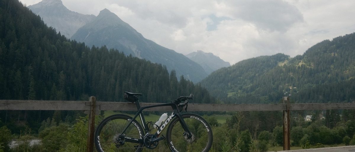 Ein Rennrad an einer hölzernen Leitplanke an einer Bergstrasse, dahinter bewaldete Hänge und hohe Gipfel im Tal.