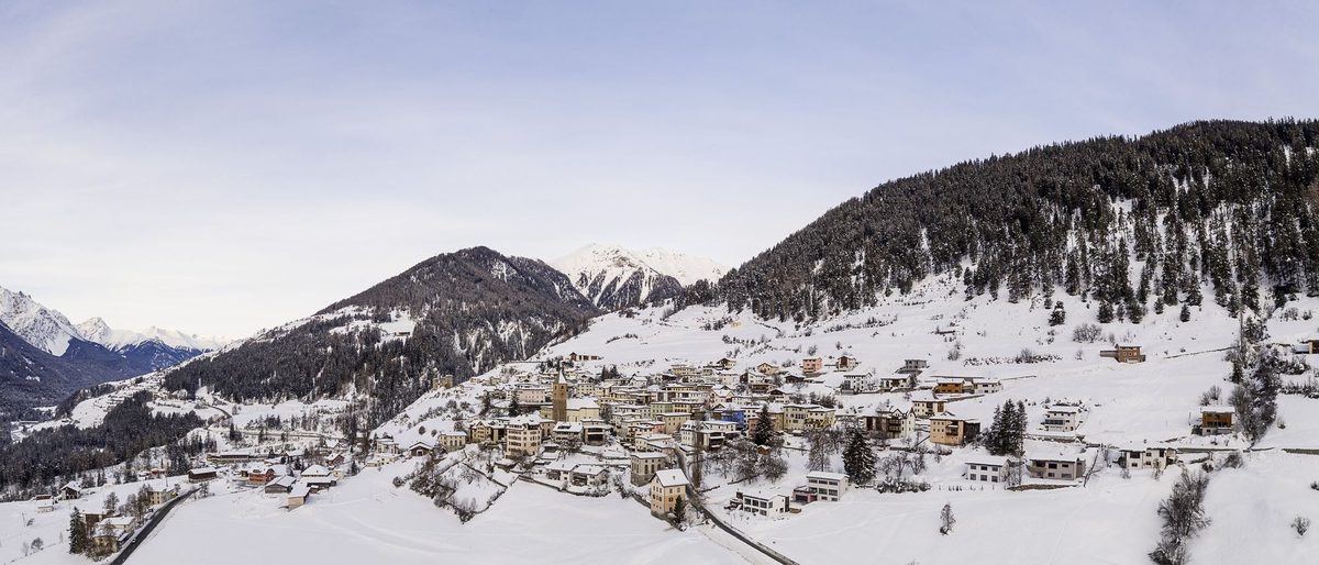 Ramosch im Schnee, von oben gesehen: die Häuser des Dorfes am sonnigen Hang, dahinter verschneite Berge.