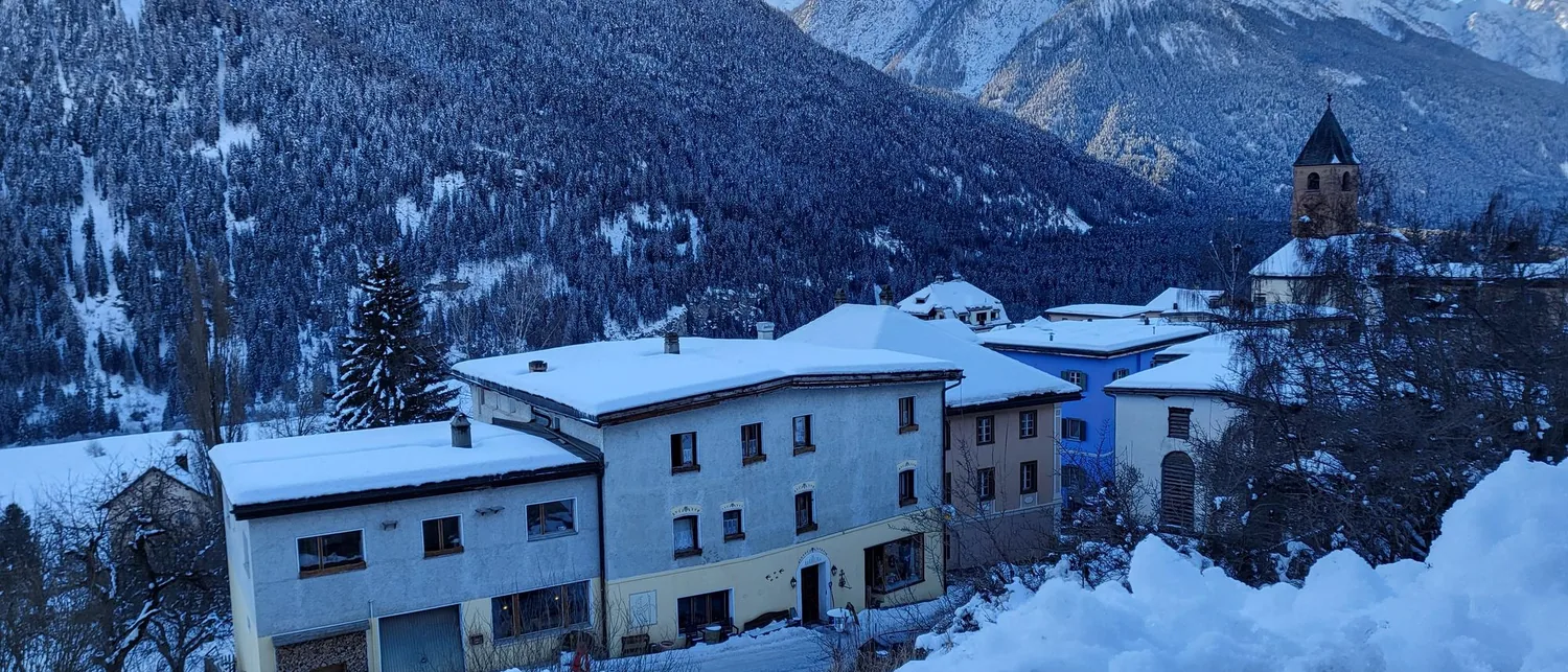 Ramosch unter Schnee zur blauen Stunde, mit dem Kirchturm über den Dächern.