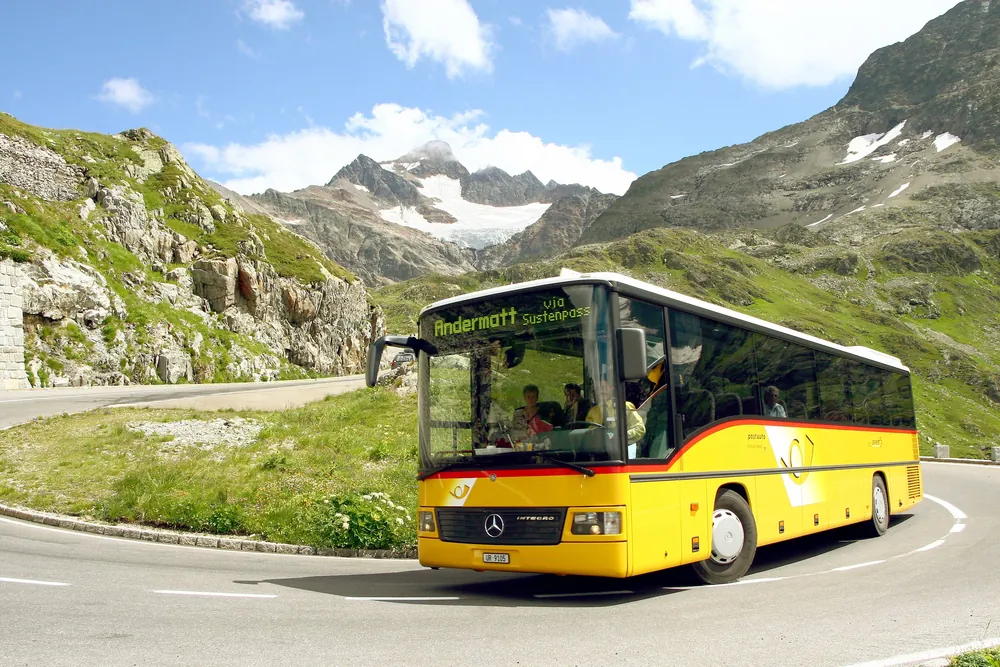 Ein gelber PostAuto-Bus auf einer kurvigen Bergstrasse, mit Felsen und verschneiten Gipfeln dahinter.