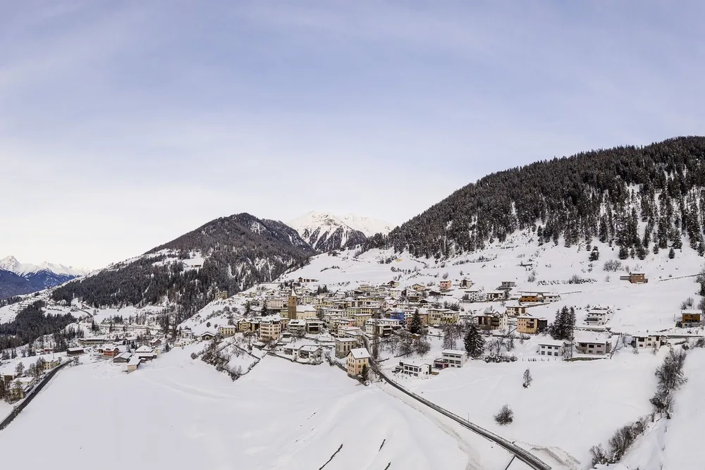 Ein Dorf unter Schnee, von oben gesehen, mit den Bergen dahinter.