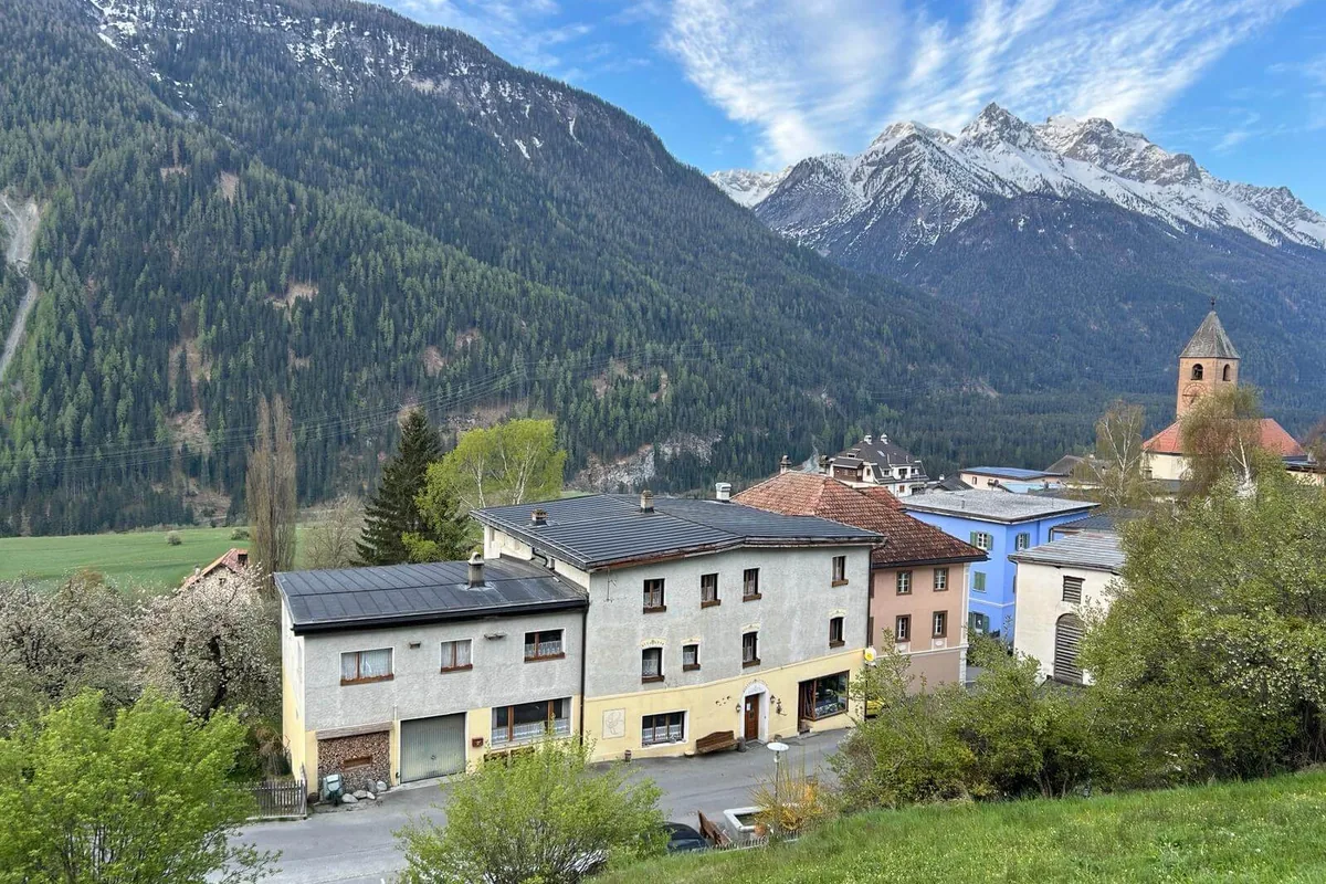 A mountain village in spring, with trees in blossom, a church tower and snow-capped peaks behind.