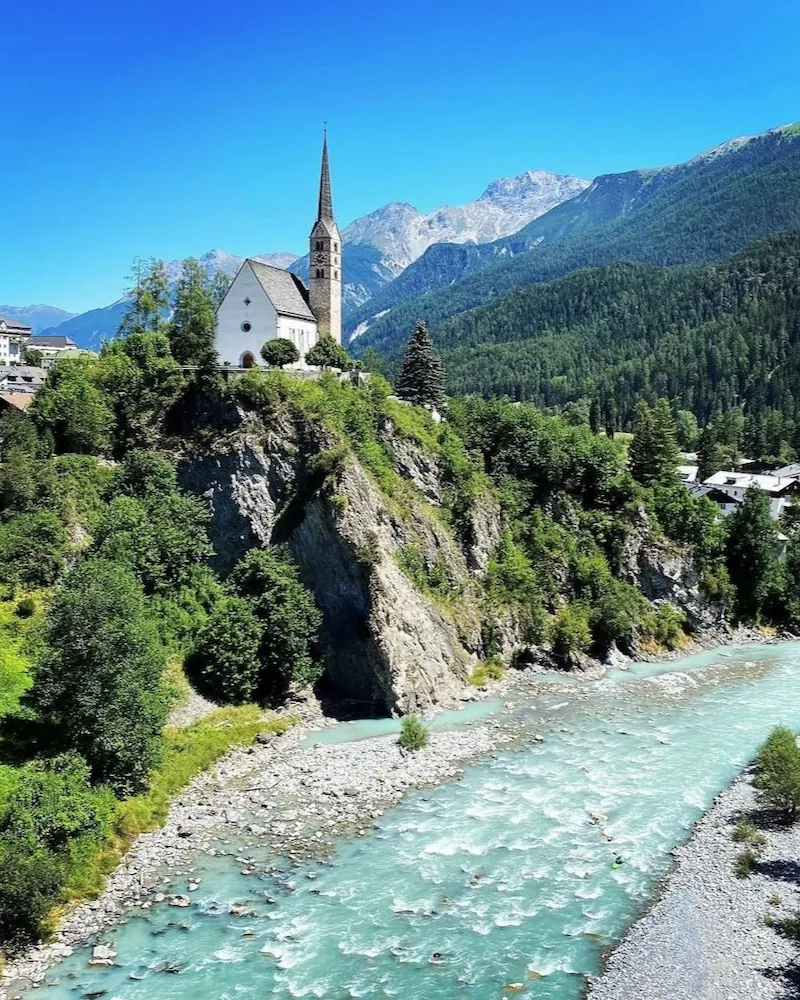 A church on the rocks above a river, with the village and the mountains behind.