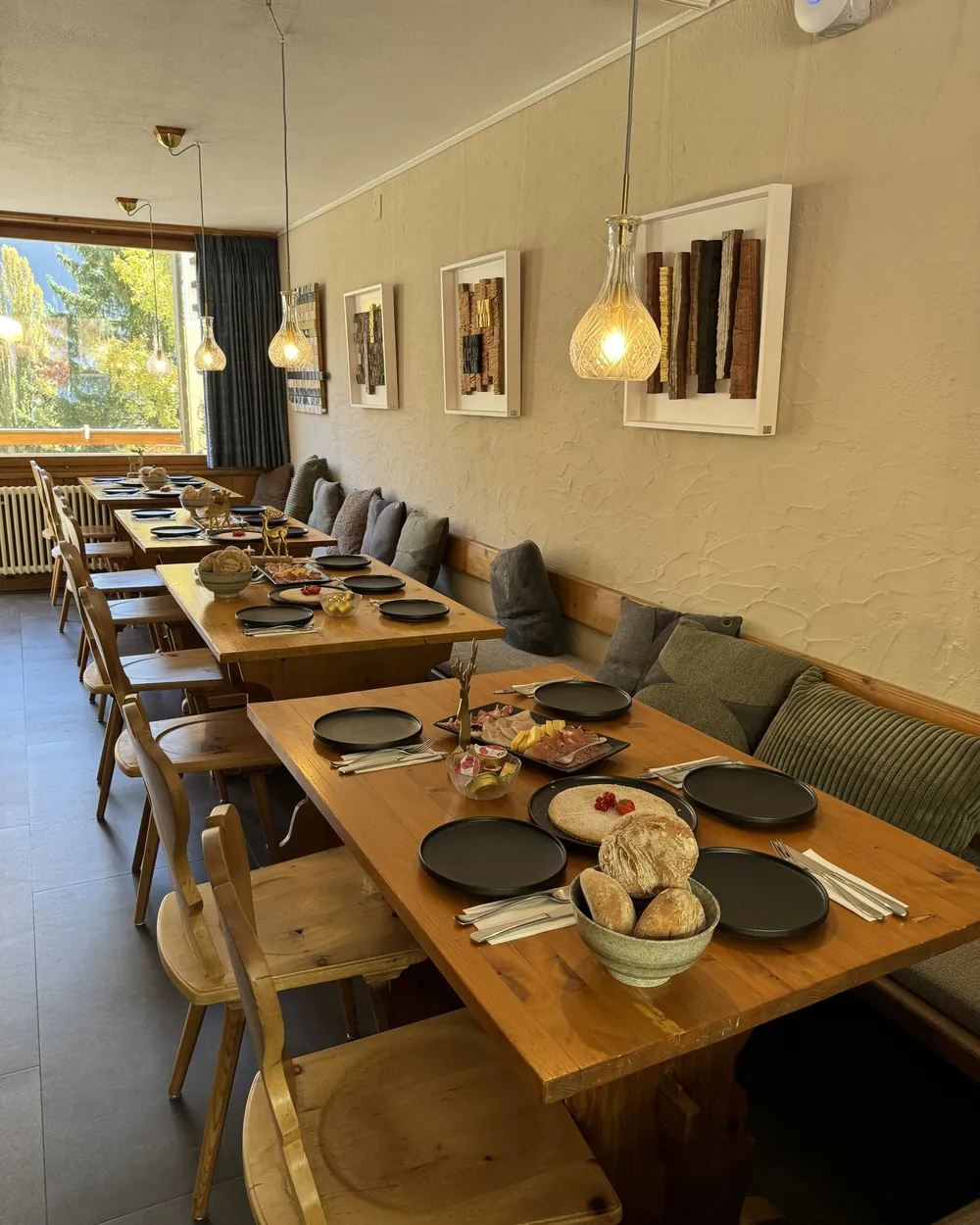 De eetzaal met gedekte houten tafels, hanglampen en ingelijste houten objecten aan de muur.