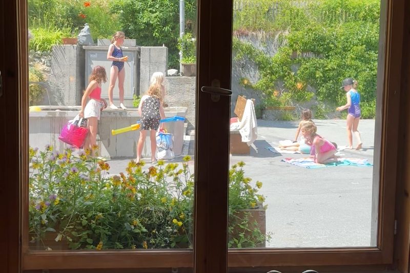 Durchs Fenster gesehen: Kinder spielen draussen auf der Strasse in der Sonne, mit einem Blumenkasten vor dem Fenster.
