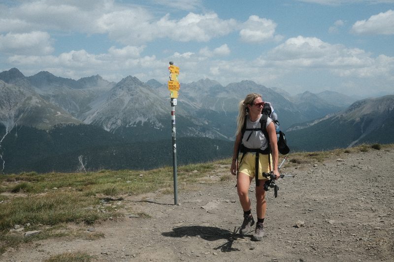 Eine Wanderin mit Rucksack an einem gelben Wegweiser, dahinter eine Bergkette.