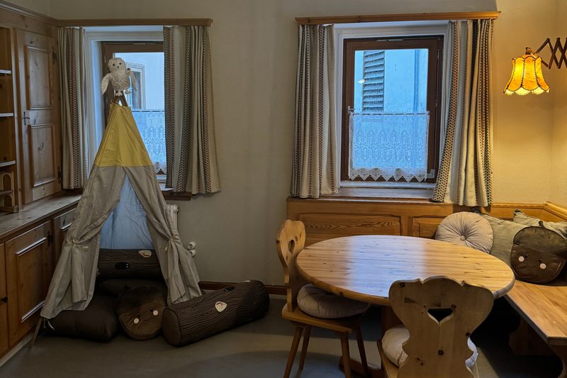 Das Spielzimmer mit einem Tipi, einem runden Holztisch mit Stühlen und Fenstern mit Vorhängen.