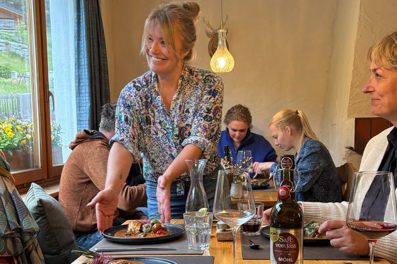 Eine Frau stellt Teller mit Essen auf einen Tisch, an dem Leute beim Essen sitzen.