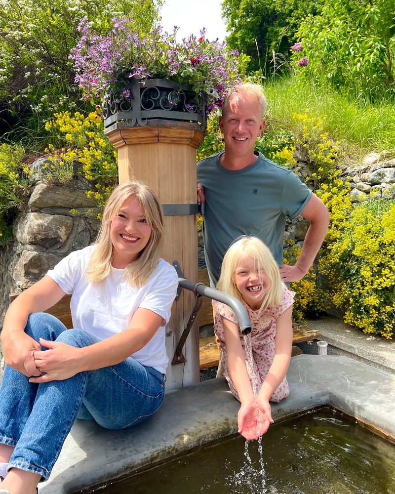 John, Ilona und Luna an einem hölzernen Brunnen zwischen den Blumen; Luna lässt sich das Wasser über die Hände laufen.
