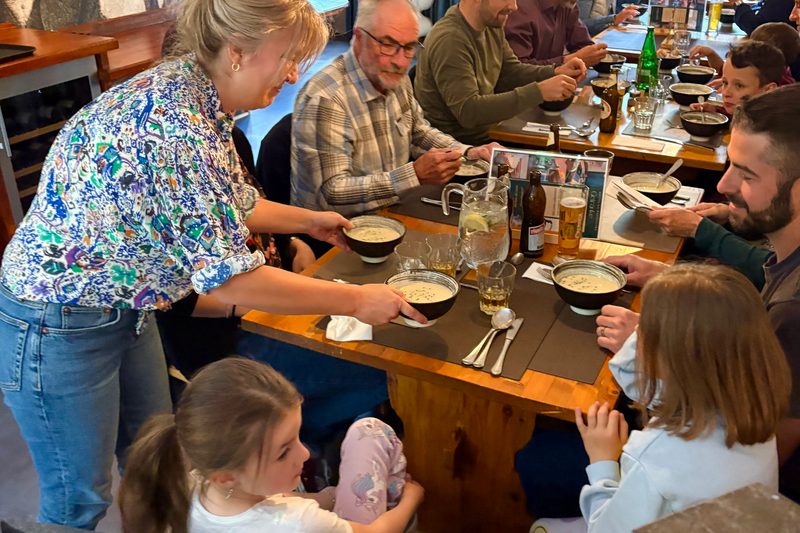 Een lange tafel vol mensen die eten, terwijl een vrouw kommen op tafel zet.