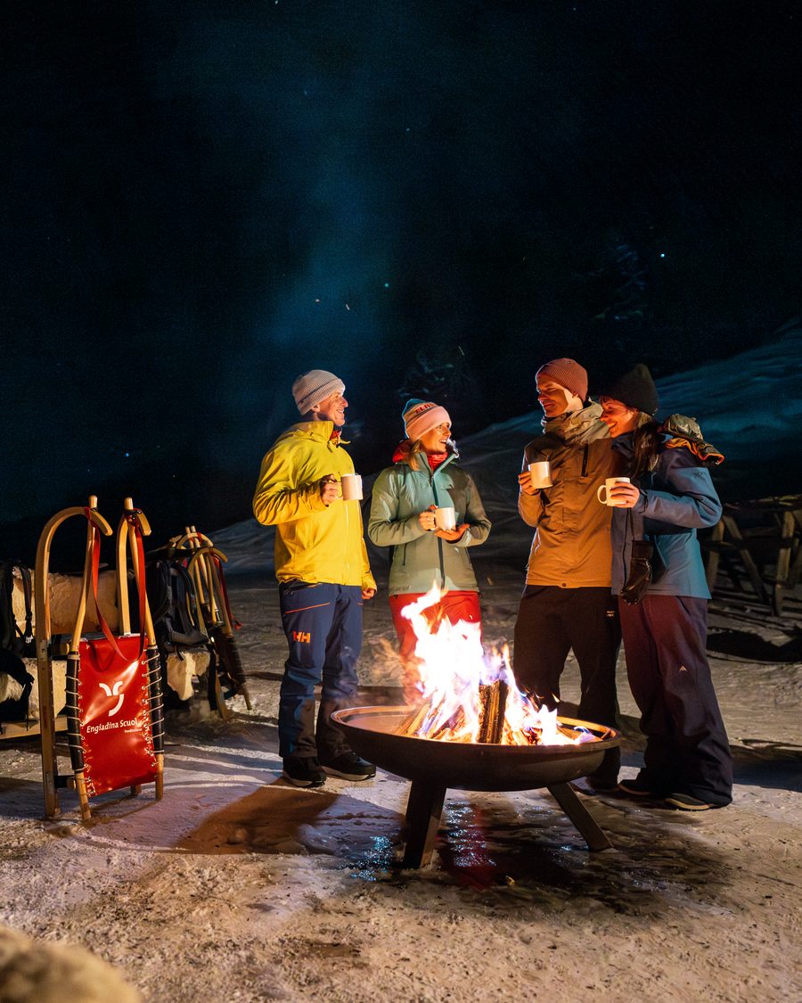 Een groep met warme drankjes rond een vuurkorf op een besneeuwd terras in de avond, met sleeën ernaast.