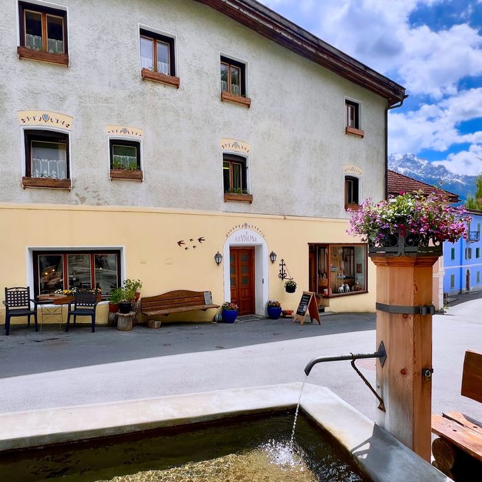 Die Vorderseite von La Randulina am Dorfplatz, mit den Sgraffito-Schwalben über den Fenstern, einer Bank neben der Tür und dem Dorfbrunnen davor.