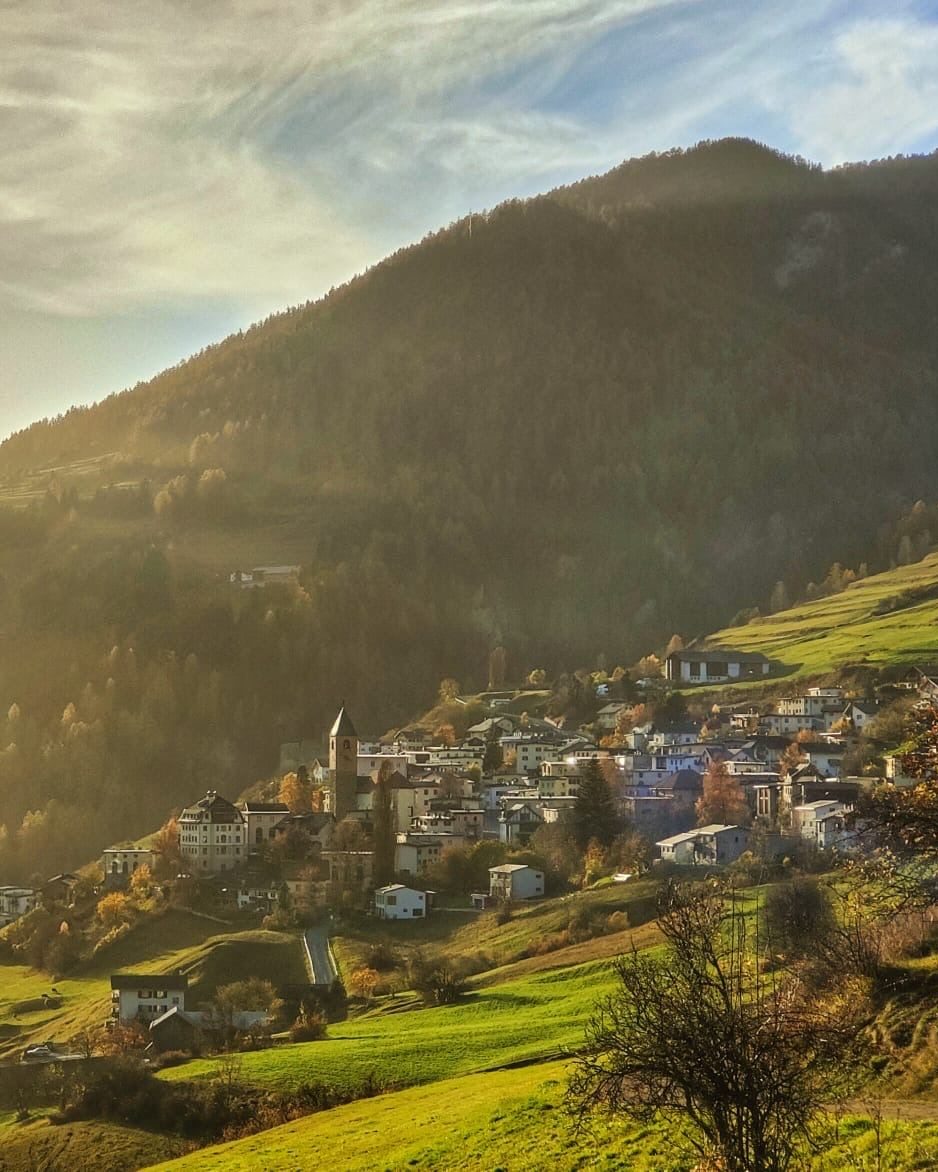 Ramosch im Herbst, vom Hang aus gesehen: der Kirchturm und die Häuser im tiefen Abendlicht, mit Herbstfarben an den Hängen.