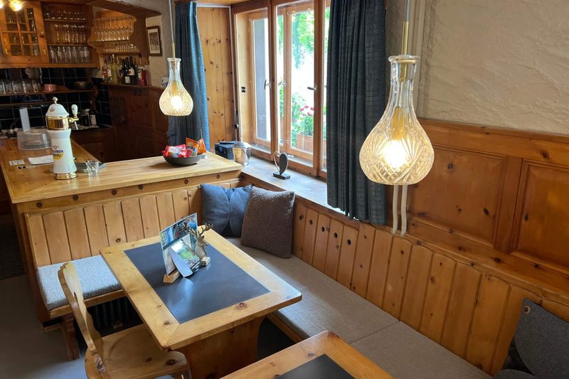 De Stube: een houten hoekbank met kussens langs het raam, een tafel, glazen hanglampen en de bar met een biertap.