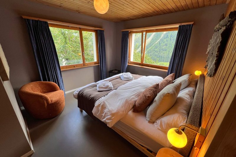 Ein Doppelbett mit aufgeschüttelten Kissen neben einem orangen Sessel, mit zwei Fenstern, die auf ein bewaldetes Tal blicken.