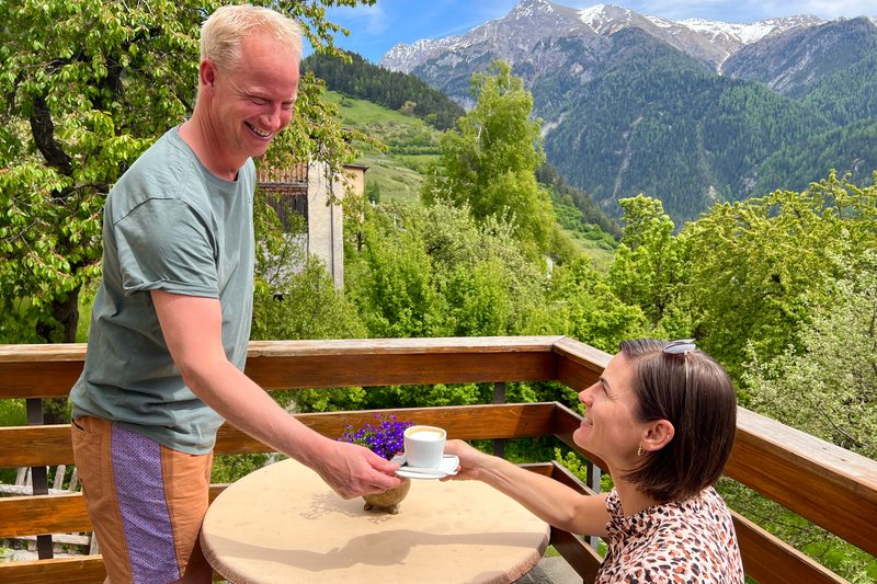 Ein Mann stellt einer Frau an einem runden Tisch draussen eine Tasse hin, mit den Bergen dahinter.