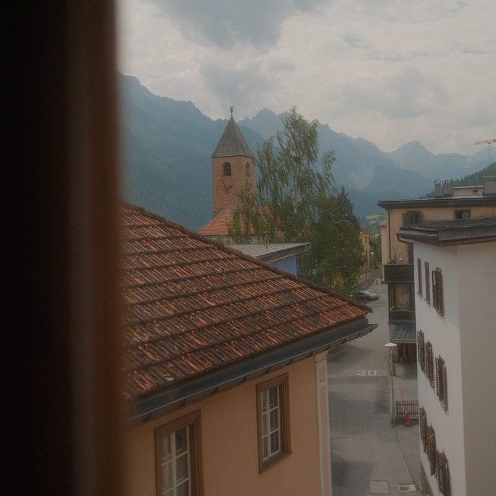 Blick aus einem Fenster auf eine Dorfstrasse mit einem Terrakottadach und einem Kirchturm, mit Bergen in der Ferne.