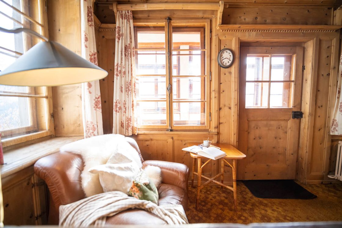 Ein Ledersessel mit einem Schaffell in einer holzverkleideten Ecke der Lodge, mit einer alten Wanduhr, einem Tischchen mit einem Buch und Licht durch die Fenster.