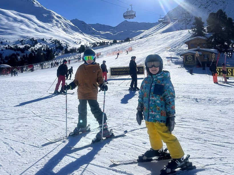 Zwei Kinder auf Ski mit Helm und Skibrille auf einer sonnigen Piste, mit einem Sessellift darüber.