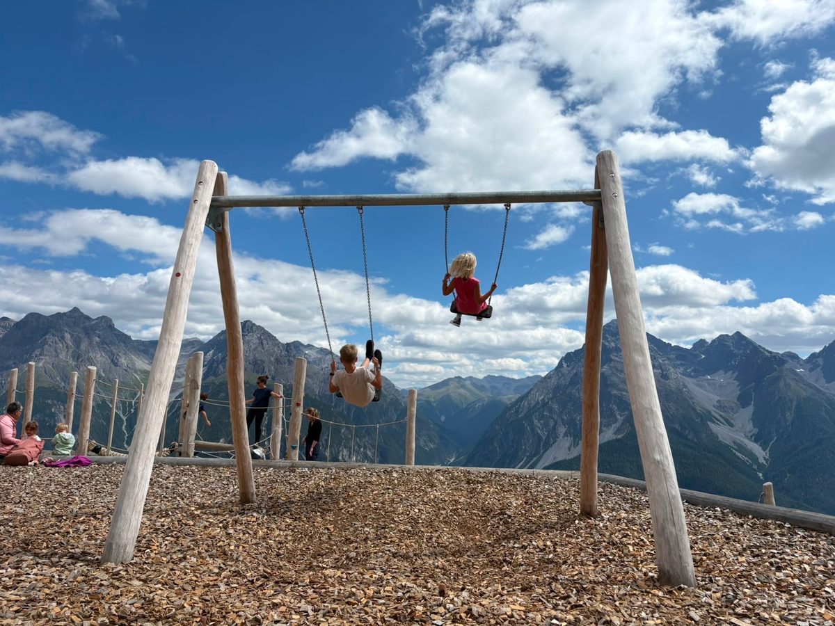 Kinder auf einer grossen Schaukel, mit Berggipfeln und Wolken dahinter.
