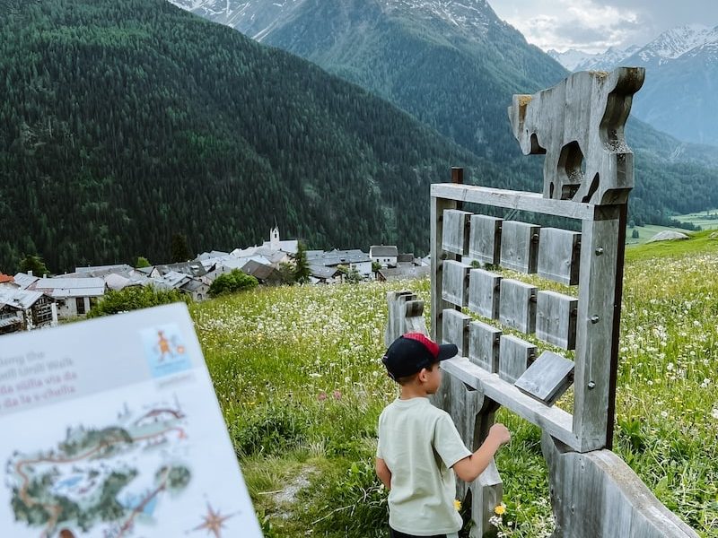 Ein Junge an einer Holztafel an einem Wanderweg, mit einer Wanderkarte im Vordergrund und einem Dorf und Bergen im Hintergrund.