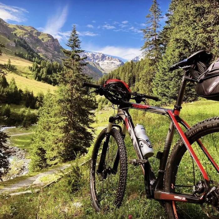Ein Mountainbike an einem Weg entlang eines Bergbachs, mit verschneiten Gipfeln über dem Tal.