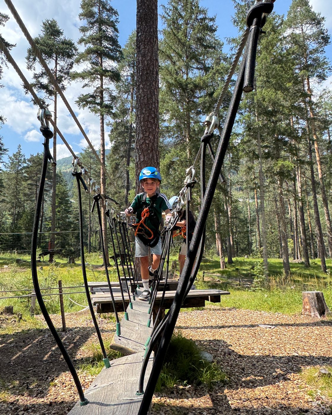 Ein Kind mit Helm geht über eine Seilbrücke auf einem Spielplatz zwischen den Kiefern.