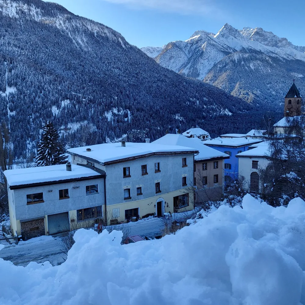 Snow-covered houses in the village, with wooded mountains and white peaks behind.