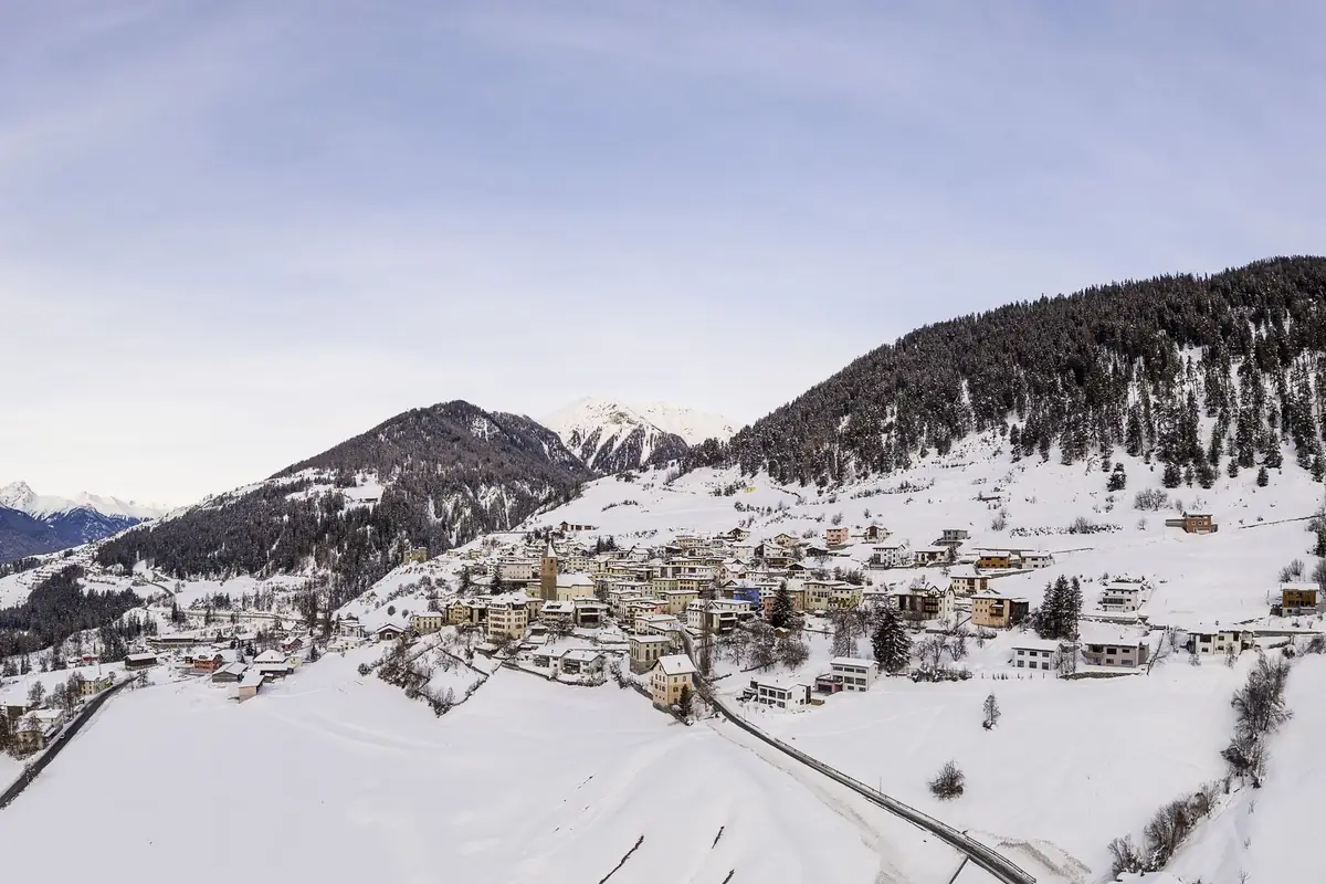 A village under snow, seen from above, with the wooded mountains behind it.