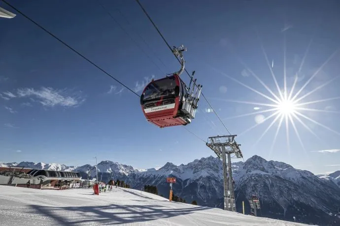 A red Motta Naluns gondola above the piste, against the sun and the mountains.