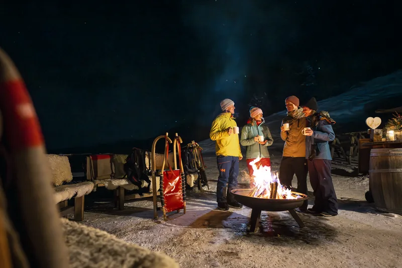 Freunde in warmen Jacken um eine Feuerschale im Schnee, unter einem Sternenhimmel.