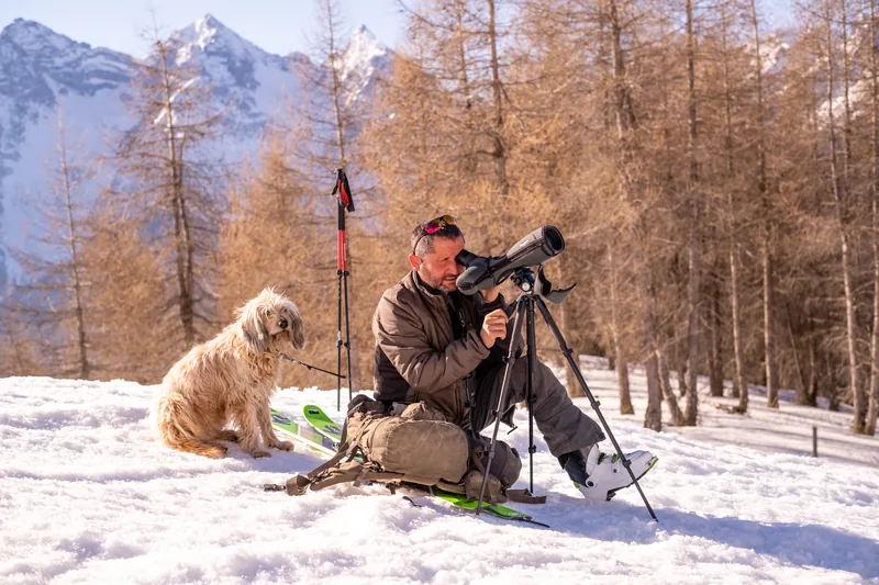 Ein Mann schaut im Schnee durch ein Fernrohr, neben ihm ein Hund, dahinter Lärchen.