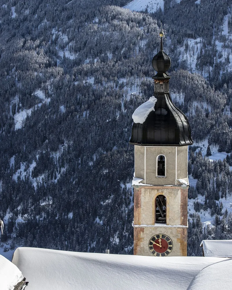 Ein Kirchturm über den verschneiten Dächern, mit bewaldeten Bergen dahinter.
