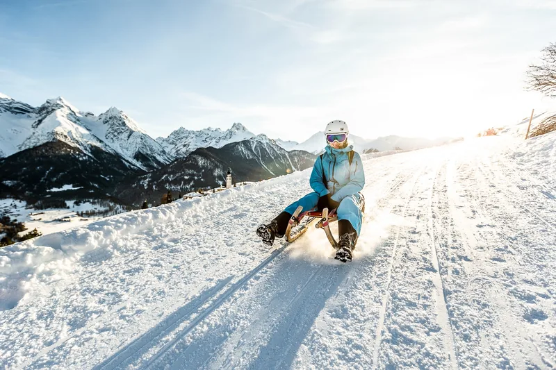 Jemand in einem blauen Skianzug, mit weissem Helm und Skibrille, auf einem Holzschlitten auf einer verschneiten Strasse, im Hintergrund verschneite Berggipfel und ein Kirchturm.