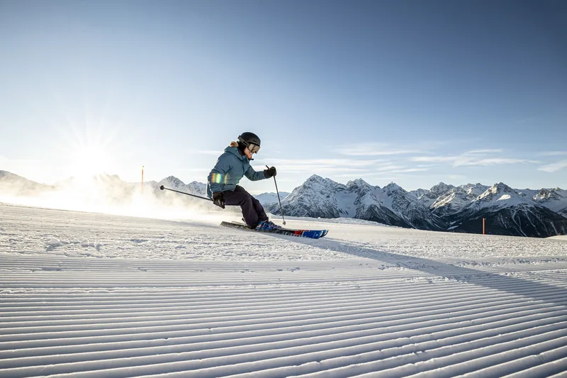 Jemand fährt auf Ski einen Bogen auf einer frisch präparierten Piste, die Sonne steht tief über den Bergen.