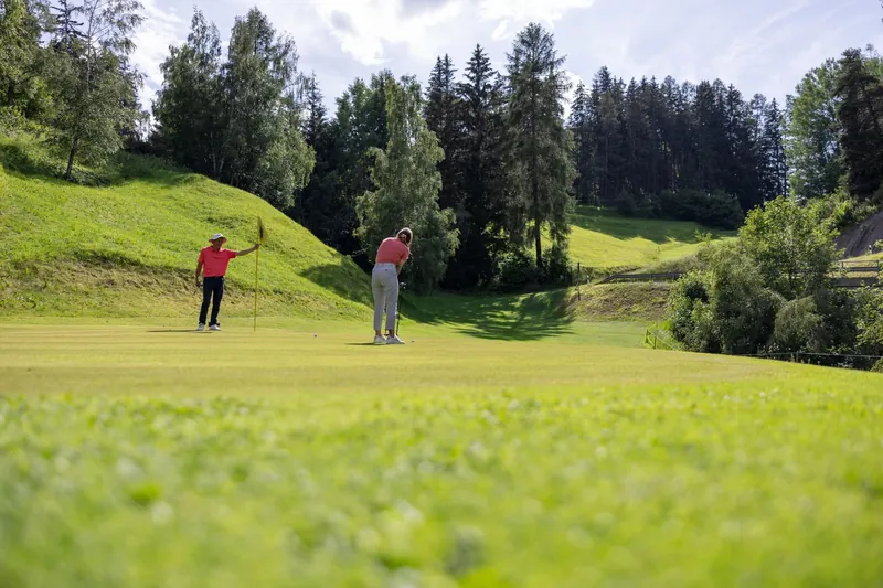 Twee golfers op een green, met dennenbossen erachter.