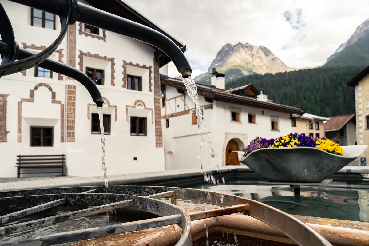 A fountain among Engadine houses in Scuol, with mountains in the background.
