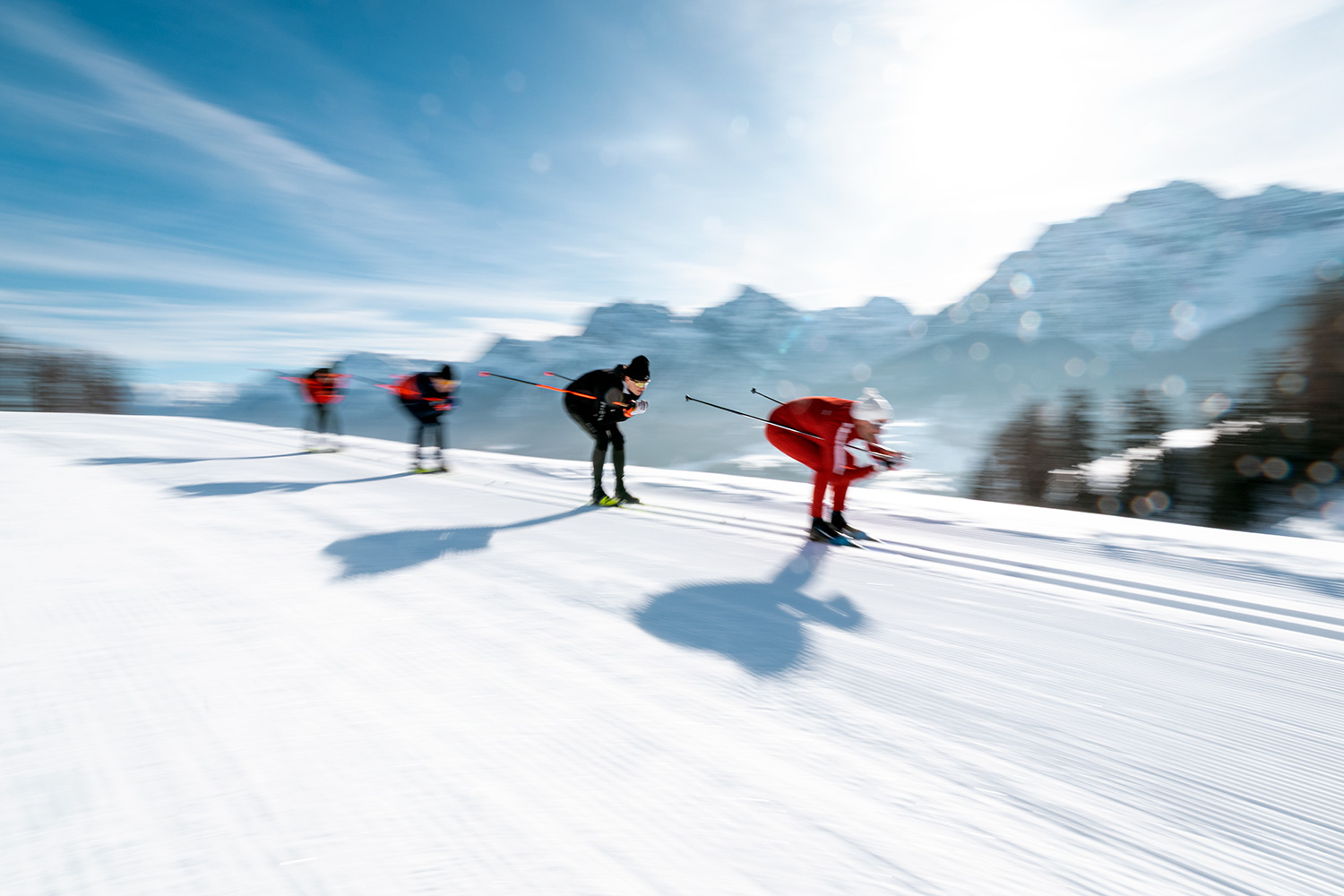 Eine Gruppe beim Langlaufen auf einer sonnigen Loipe vor verschneiten Bergen.