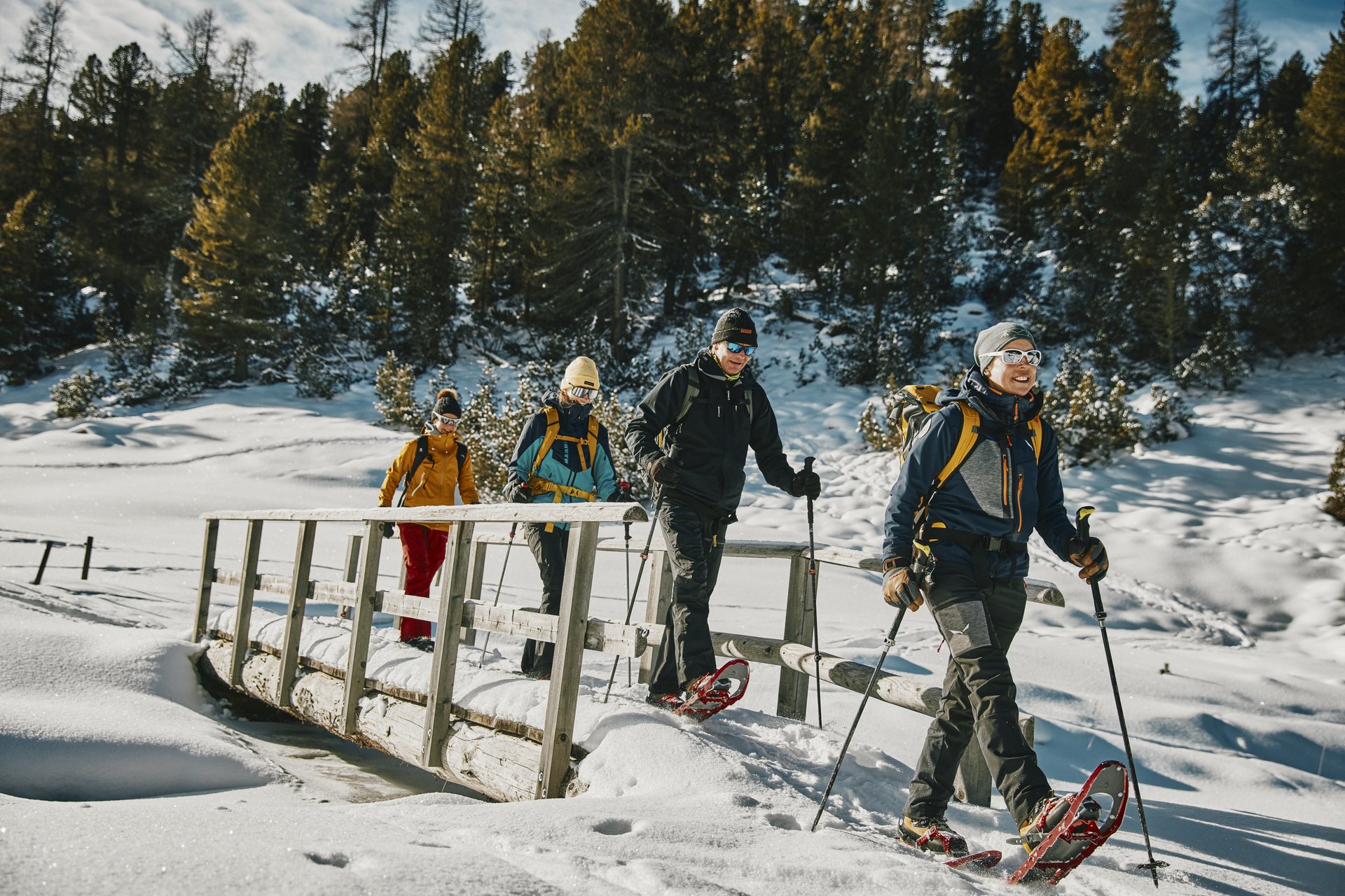 Vier Menschen mit Schneeschuhen überqueren eine Holzbrücke in einer verschneiten Berglandschaft.