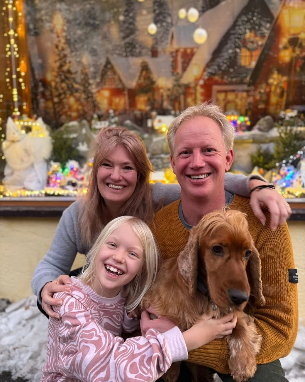 Ein lachender Mann, eine Frau und ein Mädchen mit einem Cockerspaniel, vor einer beleuchteten Weihnachtsszene.