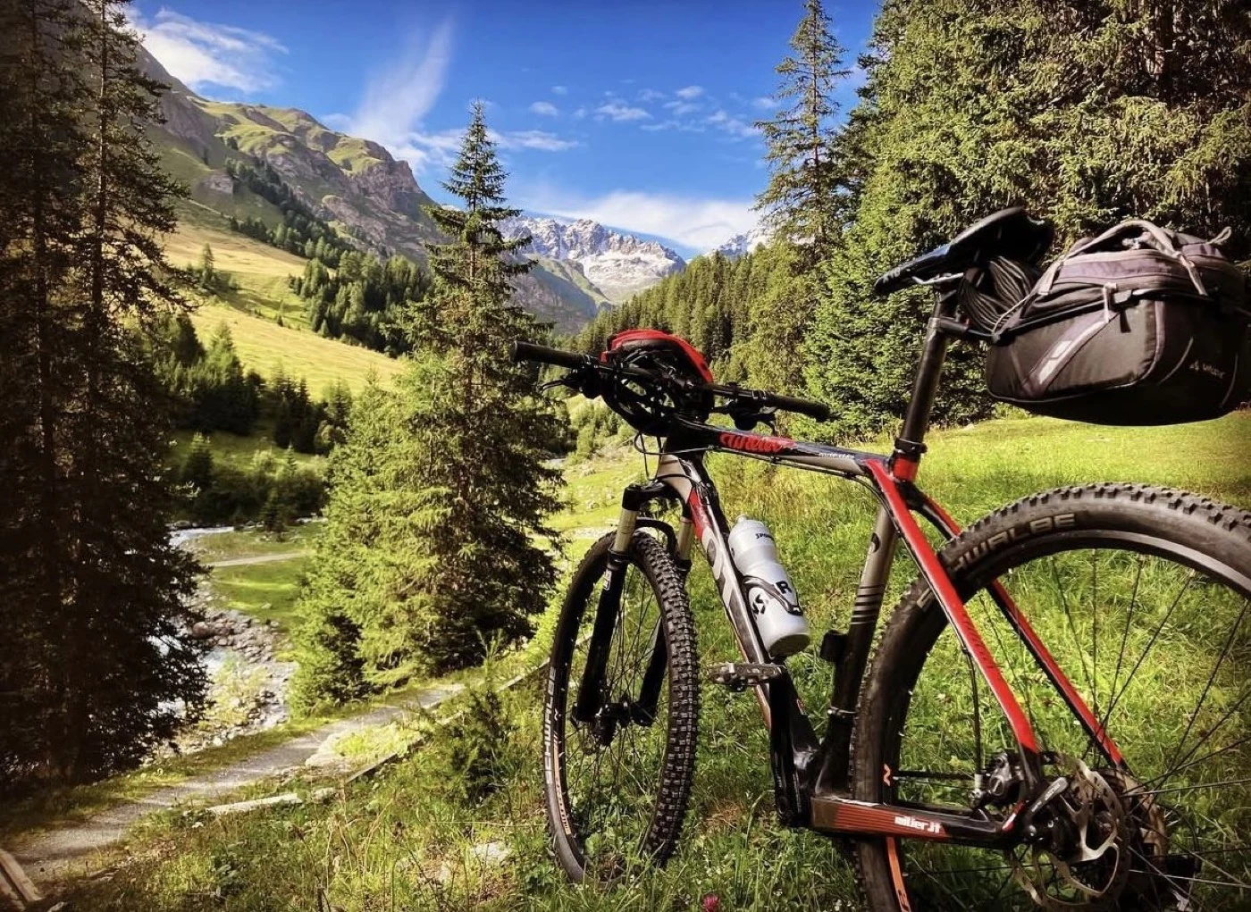 Ein Mountainbike an einem Weg entlang eines Bergbachs, mit verschneiten Gipfeln über dem Tal.